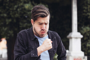 Young man hardly coughs, staying in a park, outdoors