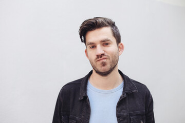 Young casual man, outdoors, in from of white wall. Man looking at the camera, mixed emotions, unsatisfied, not happy to hear news