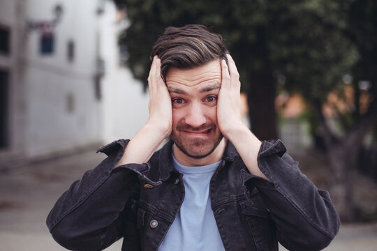 Young casual man holds his head with hands, looks scared and shocked. Concepts of getting unexpected news. Outside, outdoors, man in a park