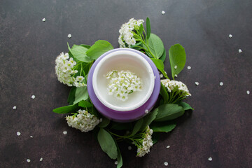 Lilac jar with white cream of delicate texture, inside which-white flowers. Spirea twigs with flowers and green leaves are lying around. Dark background 