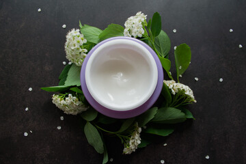 A lilac jar with a white cream of a delicate texture, around which-sprigs of spirea with white flowers. Dark background