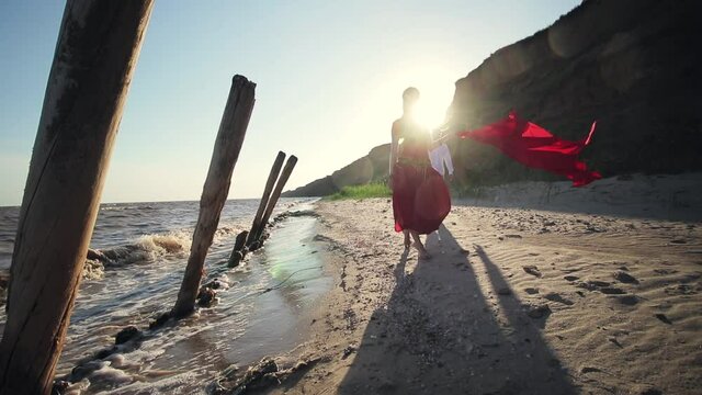 Oriental girl in a burqa and in a red caftan with belly dancing on the beach in slow motion. Red fabric flies in the wind, the sun shines brightly. Sea and sand beach