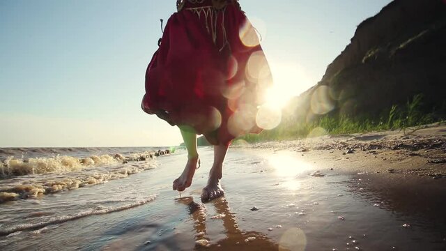 a girl in a red-eastern dress walks barefoot on wet sand on the seashore in summer in slow motion. Ocean waves, beach, and sun. Splashes of water fly into the camera