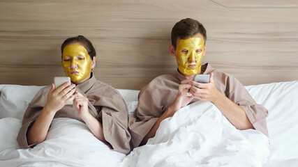 young couple with golden cosmetic face masks surfs internet with smartphones lying in hotel soft bed at self-isolation closeup