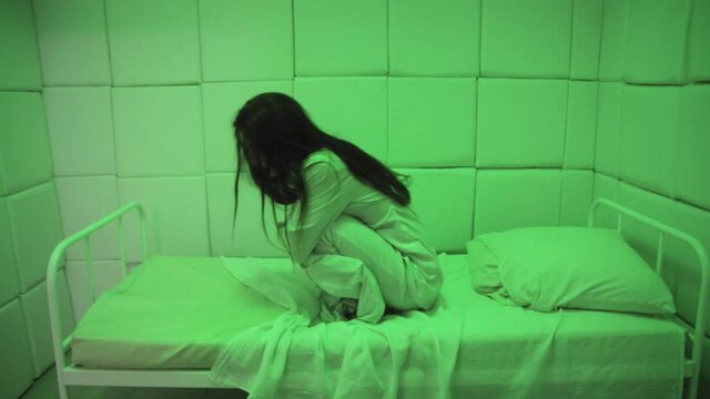 Mentally ill girl with long hair sits on a bed in a ward of a psychiatric clinic in green light. Crazy women in a straitjacket in isolation