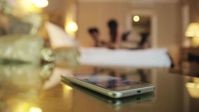 Mobile phone is on the table. On a blurred background, two lisbians girls in lingerie make love on the bed