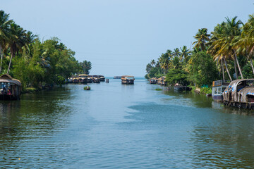 Obraz premium Small houses in a local village located next to Kerala's backwater on a bright sunny day and traditional Houseboat seen sailing through the picturesque backwaters of Allapuzza or Alleppey in Kerala 