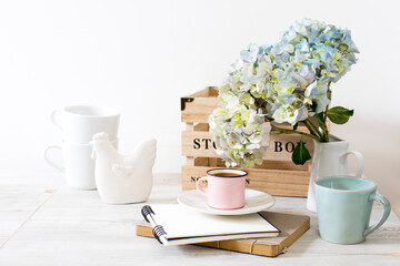 large branch with green and blue pastel hydrangea in wooden storage box, pink hydrangea in a round glass vase, a porcelain figurine of chicken, and a stack of notebooks, on a beige table.