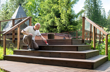 Treatment of wooden boards with protective soil, oil on wood. A man in a white T-shirt and glasses 40-50 years old, Caucasian, paints the terrace.