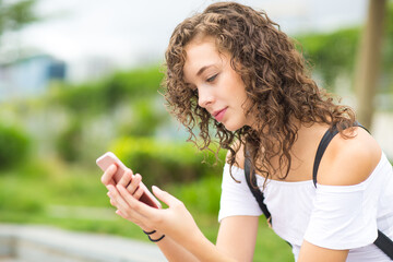 Beautiful young female teenager using her mobile phone texting in the park after school. Model with brown curly hair.