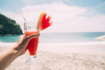 Hand with glass of cocktail with watermelon juice on the background of the ocean. Healthy red drink. 