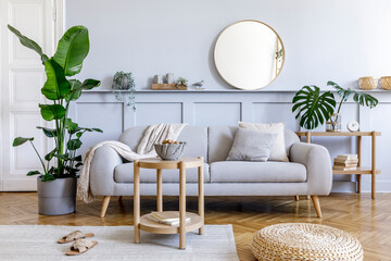 Stylish living room interior with design grey sofa, coffe table, rattan pouf, basket, shelf, mirror, tropical plants, decoration, carpet and elegant personal accessories in modern home decor.