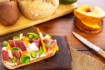 Open faced sandwich with iberico ham, parmesan and goat cheese, zucchini, pumpkin, basil and chives on sourdough bread. On a chopping board and wood, aside bread loaf, knife and pumpkin pieces.