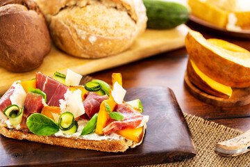 Open faced sandwich with iberico ham, parmesan and goat cheese, zucchini, pumpkin, basil and chives on sourdough bread. On a chopping board and wood, aside sourdough bread, and pumpkin pieces.