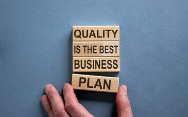 Wooden blocks form the words 'quality is the best business plan' on blue background. Male hand....