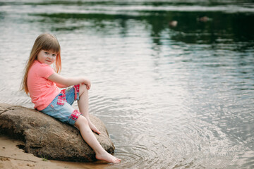 Fototapeta na wymiar Beautiful little girl is sitting on the bank of a river or sea on a stone. Looks at the camera and smiles. Copy space