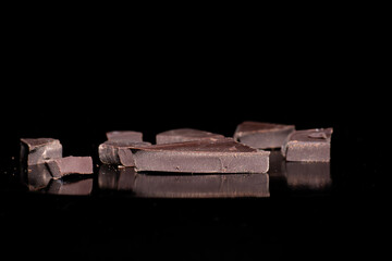 Close-up pieces of black bitter chocolate lying on a mirror surface isolated on black background,...