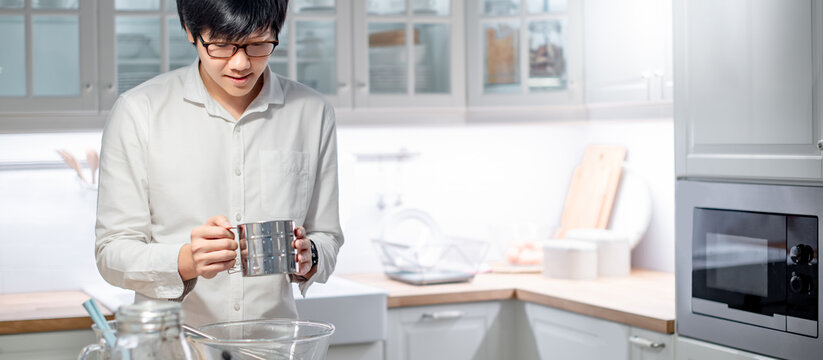 Asian man cooker holding stainless steel flour sifter preparing baking utensils on counter in the kitchen. Cooking concept