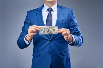 A businessman in a blue suit on a gray-blue background holds a hundred-dollar bill in front of him. The concept of generating income from participating in financial transactions.