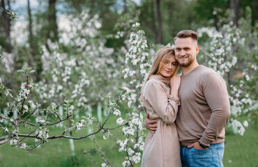 Young man and woman couple in a blooming apple orchard. Gently hold each other. Spring love story. Brown-haired woman with long hair and a guy with a haircut. Young family couple romantic spring date