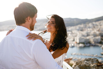 Obraz premium Back view of a beautiful just married couple, wearing in white clothing, with back panorama of city, wedding in Greece.