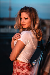 Junge hübsche Frau präsentiert Mode, posiert an einem Auto beim Sonnenuntergang in einer Stadtkulisse, Turm, Parkhaus, Lifestyle, Sommergefühlle