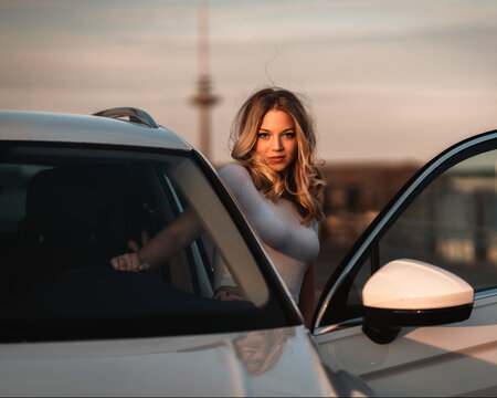 Junge hübsche Frau steig aus, posiert an einem Auto beim Sonnenuntergang in einer Stadtkulisse, Turm, Parkhaus, Lifestyle