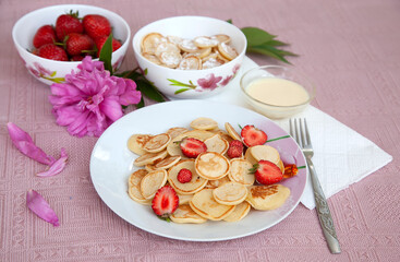 Tasty breakfast with mini pancake cereal with strawberries and sugar powder. Mini pancake cereal is new trendy food for activities during Covid-19 lockdown. 
pink tablecloth and peony