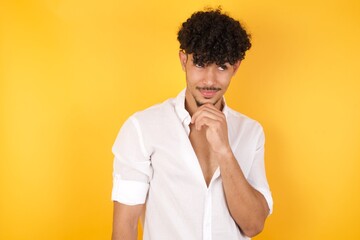 Dreamy male with pleasant expression, wearing casual clothes, looks sideways, keeps hand under chin, thinks about something pleasant, poses against gray background.