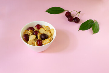 A white bowl of cereal with a tiny pancake with cherries, berries and green leaves in the background on a pink background. Copy space. Trendy food concept.