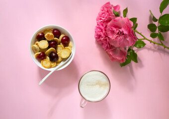 A white bowl of cereal with a tiny pancake with cherries, a cup of cappuccino and flowers on a pink background. Top view. Trendy food concept.
