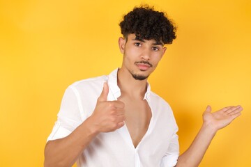 Young happy cheerful man showing thumb up and pointing with the other hand while standing indoors. Good Job. Wearing casual clothes.