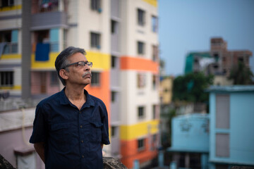 Body portrait of an An old/aged Indian Bengali man in blue shirt is standing on a rooftop under the open sky in front of a urban landscape. Indian lifestyle and seniors