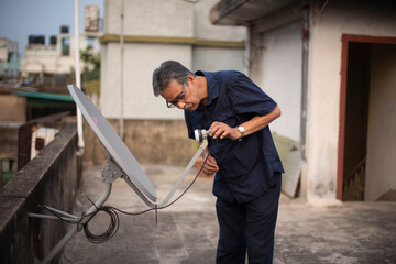 An old/aged Indian Bengali man in blue shirt is trying to fix a dish antenna standing on a rooftop under the open sky. Indian lifestyle and seniors