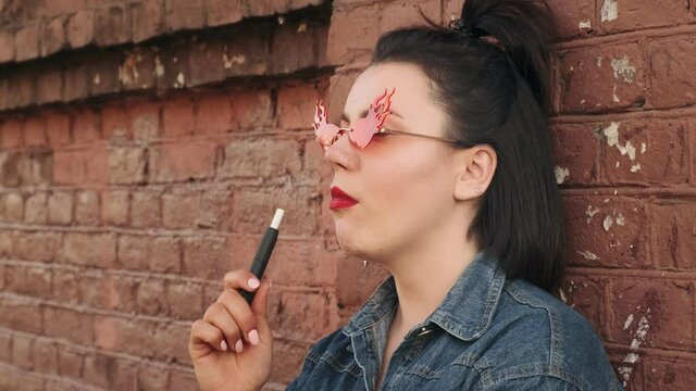 Stylish young woman vape gadget cigarette, puffs on a vape pen device. Electronic cigarette outdoors. Smoking tobacco stick, smoke.