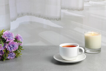 a white cup of red tea and a glass of candle are on the grey table with bouquet of flowers in the white living room near the white curtain