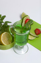 Green lemonade in a glass. Decorated with lime and strawberries. With sprigs of mint. Near lemon and mint.