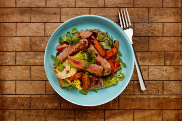 A delicious bowl of BBQ rump steak strips, sweet potatoe chips and vegetables, in a light blue bowl shot on a wooden background and fork