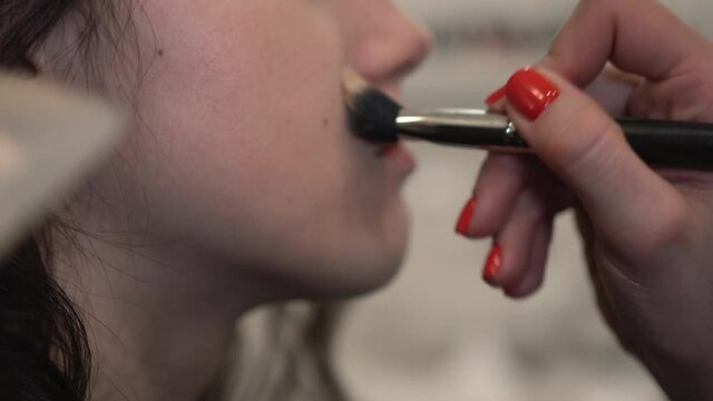 Professional makeup artist apply light liquid foundation on client&rsquo;s skin with concealer brush to even out face skin tone. Visagiste works on natural makeup for a young brunette woman with lush curls.