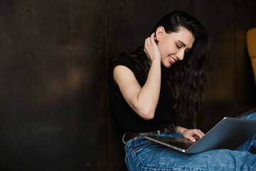 Photo of young brunette woman sitting on floor and using laptop