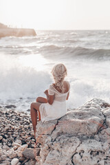 Lonely beautiful romantic girl with blonde wavy hair in white sexy dress looking at the sunset and storm on the sea and dreaming. Freedom concept