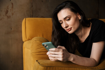 Photo of young woman lying on couch in apartment and holding cellphone