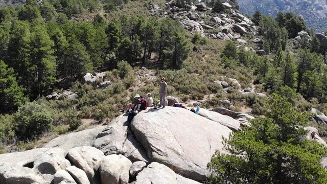 Group of friends explorers stands on summit, talks, enjoys a view of mountain ranges, valleys; silhouettes of three hikers on sunset; aerial view, drone flies up in Navacerrada, Madrid