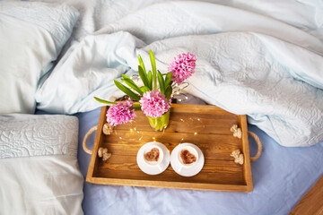 Fototapeta na wymiar A bouquet of beautiful pink hyacinths is standing in a vase on a tray on which are two cups of cappuccino with a heart pattern, the concept of a surprise in bed, a beautiful sunny morning