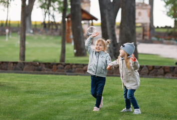 Naklejka na ściany i meble Two cute girls run on green grass holding hands and catch soap bubbles.