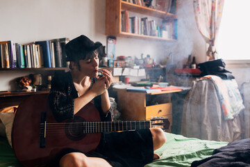 Relaxed woman in stylish clothes and hat sitting on bed with crossed legs and smoking cigarette with closed eyes