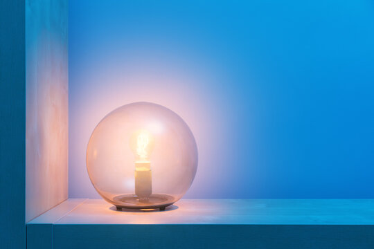 Stylish round lamp with glowing yellow light inside of glass ball locating on shelf in dark room with blue walls