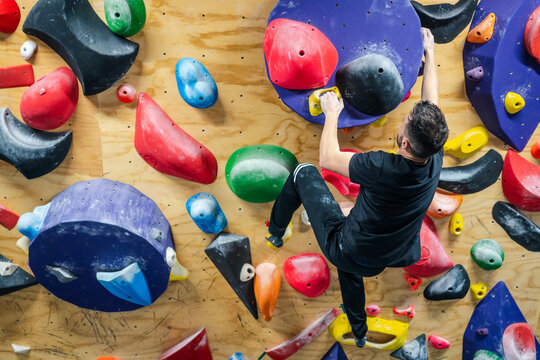 Back view of unrecognizable strong male athlete in sportswear climbing on colorful wall during workout in modern climbing center