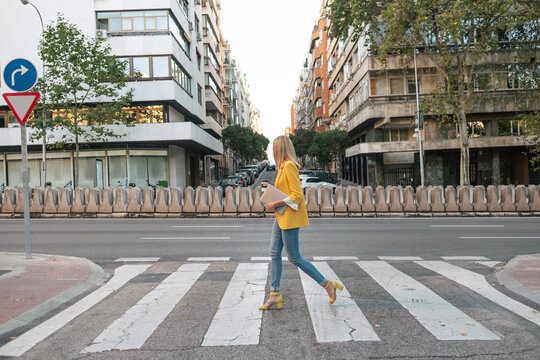 Side view of trendy slim blond female in elegant vivid yellow jacket and jeans with digital tablet walking alone along pedestrian crossing against exteriors of residential multistory buildings and cars parked on street in downtown