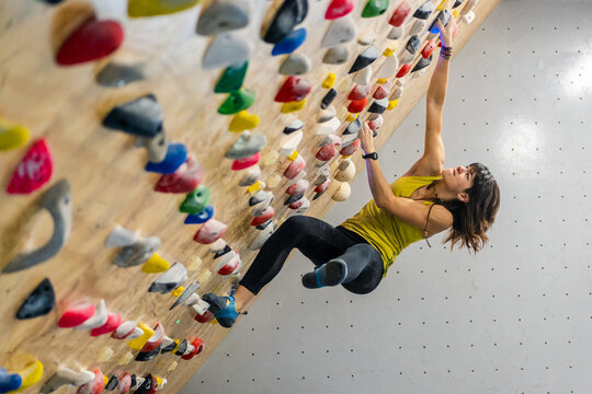Side view from below of focused woman in sportswear hanging on steep wall above mats in modern climbing center